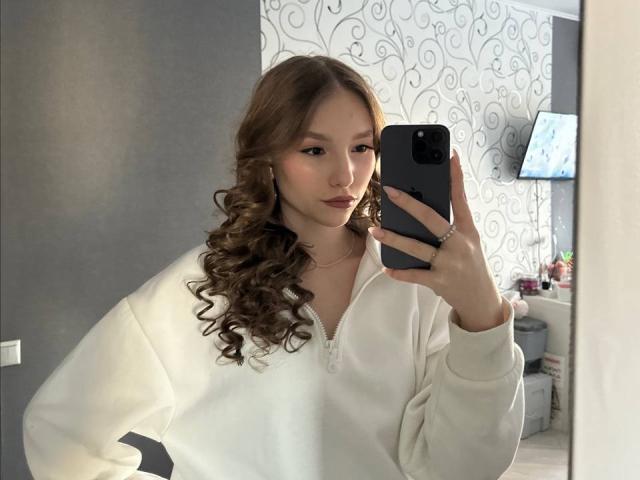 TericaSarley met krullend bruin haar, een witte zip-up hoodie, die een zwarte telefoon vasthoudt, een spiegel-selfie maakt, subtiele make-up draagt, een kleine neusring heeft, een ring om haar rechterhand draagt en een kalme uitdrukking heeft..