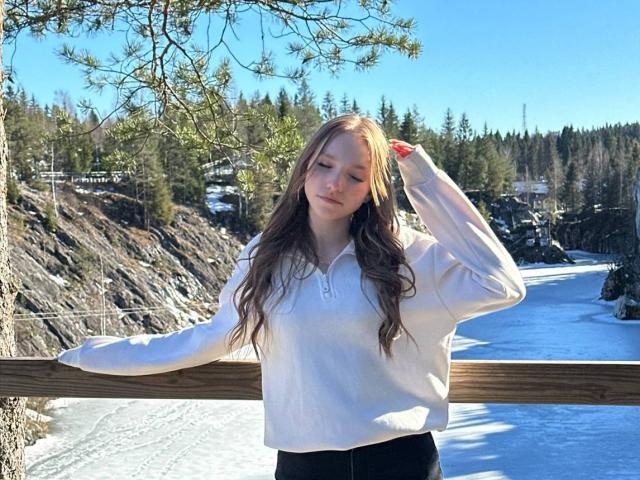 TericaSarley met lang bruin haar, een wit shirt met lange mouwen, zwarte broek, staand met haar arm op een houten leuning, hand in haar haar, helderblauwe lucht, besneeerd landschap, een dennenboom op de voorgrond..