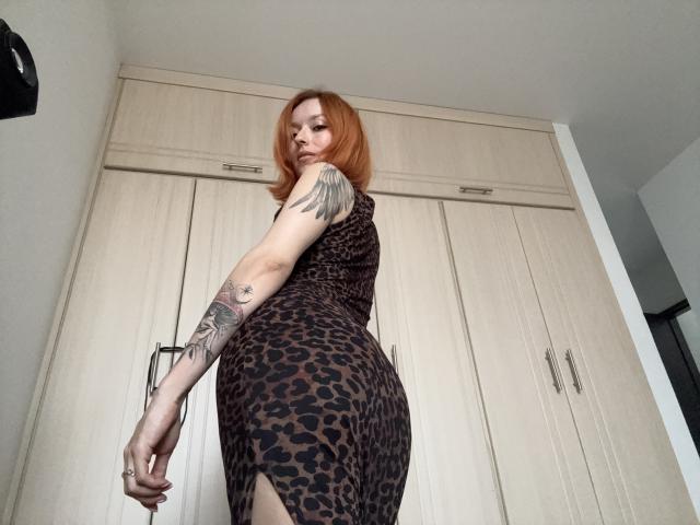 Sarah Rousen, con cabello rojo, vestida con un vestido de estampado de leopardo, mostrando su trasero y un tatuaje en su brazo derecho, mirando por encima del hombro en una habitación con armarios de madera..