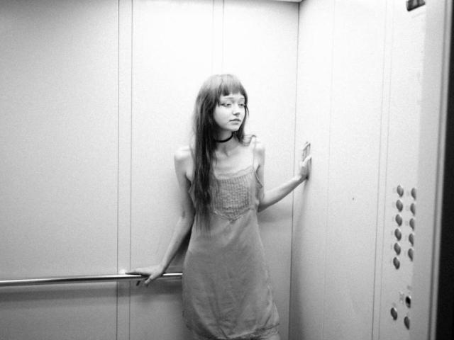 MeiWasinger, con cabello largo y lacio, vestida con un vestido transparente, está de pie en un ascensor, tocando la pared con una mano, mirando hacia un lado, con un collar negro alrededor de su cuello..