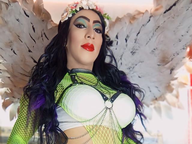KattyDirty with big tits, wearing a green fishnet top, white bra, red lipstick, flower crown, and white angel wings, has purple hair and bold green eye makeup.