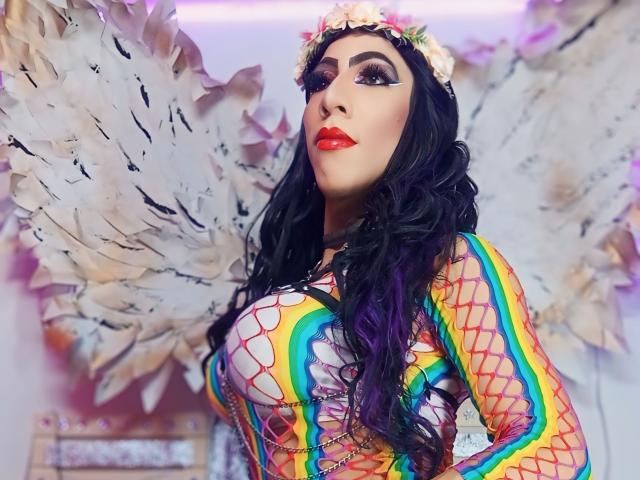 KattyDirty mit großen Brüsten, trägt ein farbenfrohes Netzhemdkleid, einen Blumenkranz und Engelsflügel, hat lila Haare, roten Lippenstift und auffälliges Make-up und blickt mit einem selbstbewussten Ausdruck nach oben..
