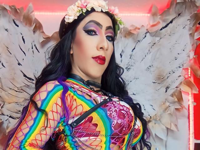 KattyDirty met een kleurrijke regenboogjurk, grote borsten, witte engelenvleugels, een bloemenhoofdband, paars oogschaduw, rode lippenstift, zwart haar en een sexy, zelfverzekerde uitdrukking..