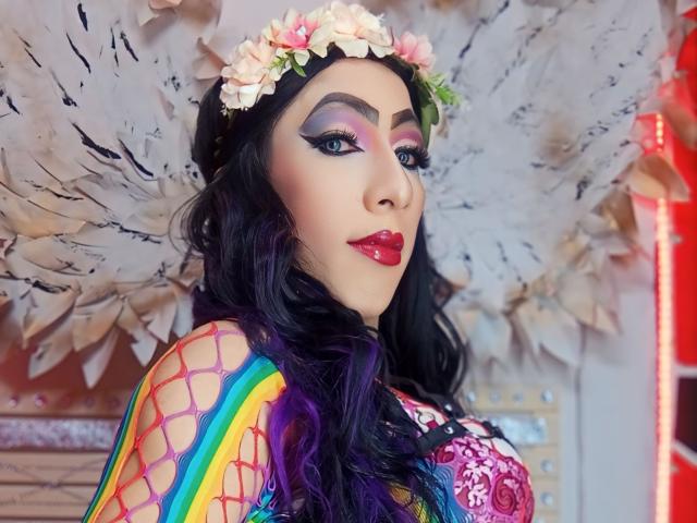 KattyDirty, con cabello morado oscuro, lleva una corona de flores, un top de malla de colores y lápiz labial rojo. Tiene ojos azules, sombra de ojos llamativa y un aspecto lindo, sexy y femenino..