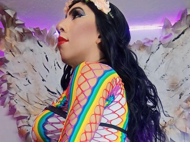 KattyDirty, com longos cabelos pretos, vestindo uma blusa de rede colorida, uma tiara de flores rosa e batom vermelho, mostrando os seios, em um perfil lateral, com asas brancas e emplumadas..