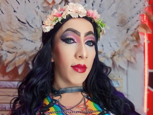 KattyDirty, avec ses cheveux violet foncé, son couronnement de fleurs, son fard à paupières rose et violet, son rouge à lèvres rouge, son collier en chaîne, son haut arc-en-ciel et ses yeux bleus, regardant directement l'appareil photo..