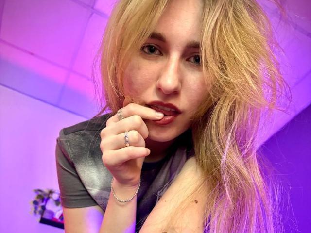 GingerLiam with long blonde hair, biting her finger, wearing a gray shirt, purple-lit, showing her tits with a cute expression, ring on her finger, bracelet on her wrist.