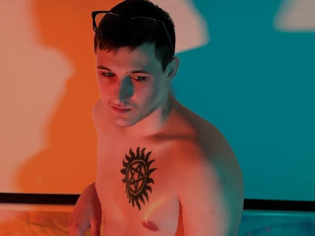 GingerLiam, shirtless with a sun tattoo and pentagram on his chest, wearing black glasses, looking down, lit with orange and blue light, short brown hair.
