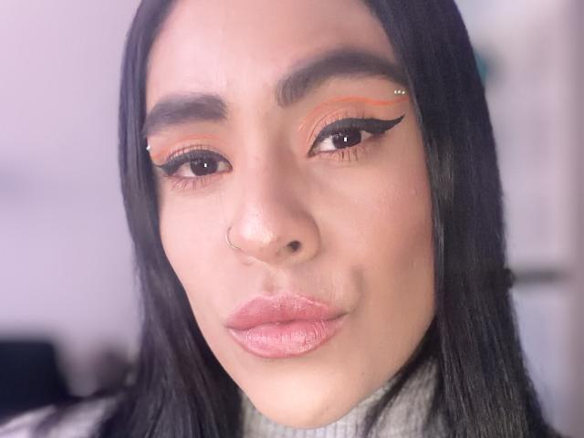 StellaBeckker's face, with long black hair, orange eyeliner, and black winged eyelashes, has a small nose ring, full pink lips, and a neutral expression in a soft-lit photo.