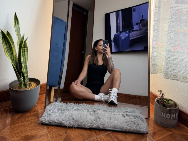 ThaliaAres sitting on wooden floor, wearing black tank top, shorts, white socks, and sneakers, taking mirror selfie, showing tattoos on right arm, with snake plant and gray rug.