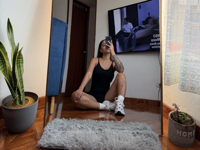 ThaliaAres with tattoos, wearing a black tank top and white sneakers, sitting on wooden floor, taking a mirror selfie, showing her ass, with a snake plant to the left.