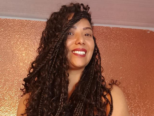 KenyaKarath, con dreadlocks neri e ricci, sorride con rossetto rosso, mostrando denti bianchi, indossa una maglietta senza maniche, ha la pelle marrone, ed è ritratta di fronte a un muro testurizzato di colore rame..
