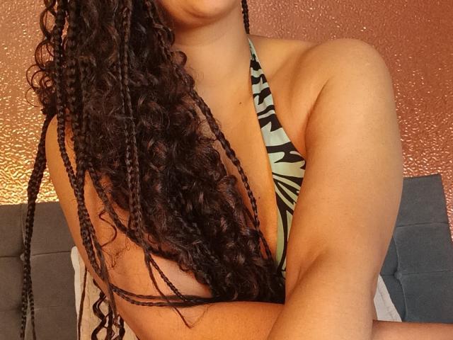 KenyaKarath with curly braids, wearing a zebra-patterned bikini, has her back to the camera, showing her bare ass and arm, against a textured, orange wall.