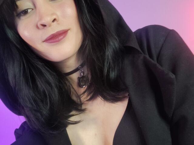 IssisDiosa with black hair, black hoodie, and black choker with ring, showing cleavage, pink and purple background, smirking with red lipstick, light skin.