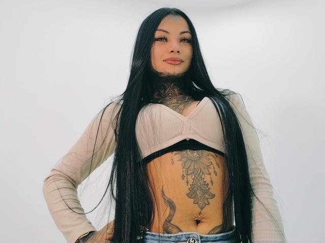 LisaMont, com cabelos pretos longos, pele bronzeada, tatuagens no peito e abdômen, sutiã rosa, cardigan bege e jeans azuis, olhando para a câmera com um leve sorriso..