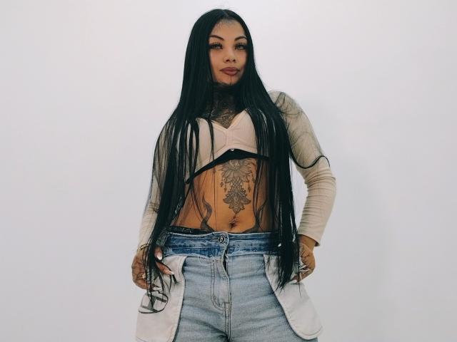 LisaMont met lang zwart haar, bruine huid, een getatoeëerde buik en een onthullend beige top, draagt blauwe jeans en een witte jas, met haar handen op haar heupen..