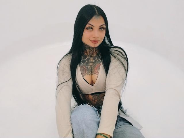LisaMont, con lunghi capelli neri, tatuaggi sul petto e sul collo, indossa una maglietta bianca con un profondo scollo, jeans blu e sorride, seduta contro un muro bianco..