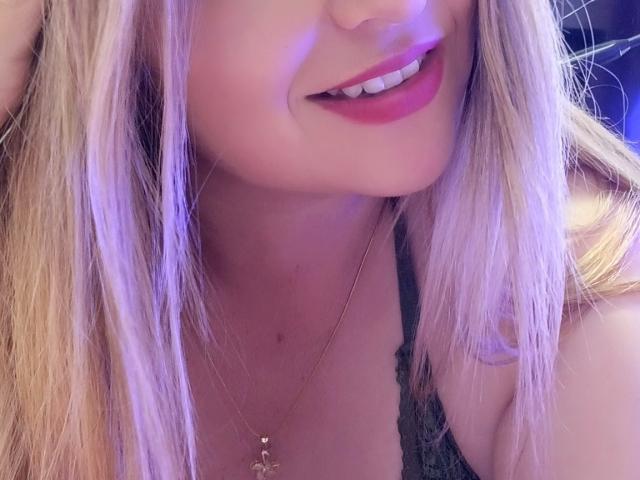 NatalyHami, con i capelli biondi, sorridente, che mostra un rossetto rosa, indossa una maglia nera in pizzo, una collana dorata con un piccolo ciondolo e un leggero riflesso viola sulla pelle..