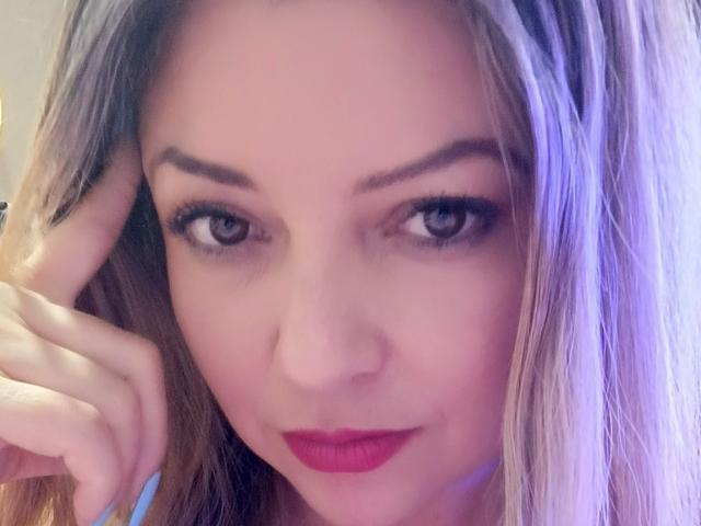 Il viso di NatalyHami, con occhi azzurri, rossetto rosso e capelli castano chiaro, che tocca la testa con una mano, mostrando il suo aspetto carino e sensuale..