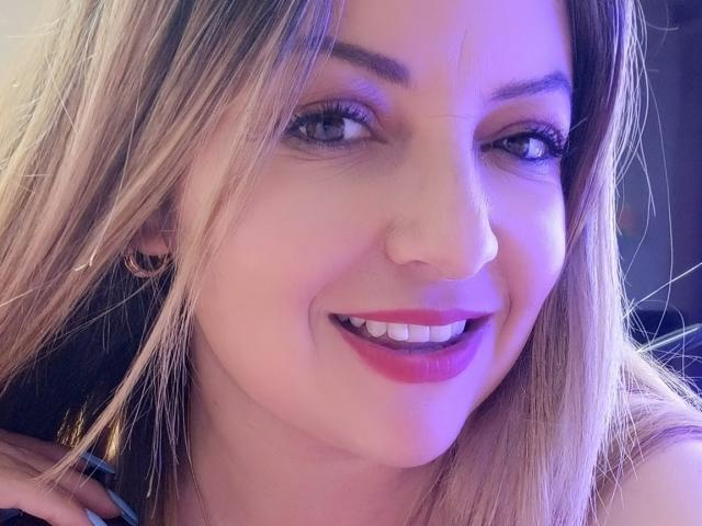 NatalyHami ha i capelli biondi, gli occhi azzurri e il rossetto rosa, sorride mostrando i denti bianchi, indossa piccoli orecchini neri e ha un naso carino con una leggera sfumatura rosa-viola..