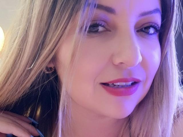 NatalyHami's face with blonde hair, purple lipstick, and a slight smile, showing her teeth. She has blue eyes, subtle makeup, and is looking slightly to the left.