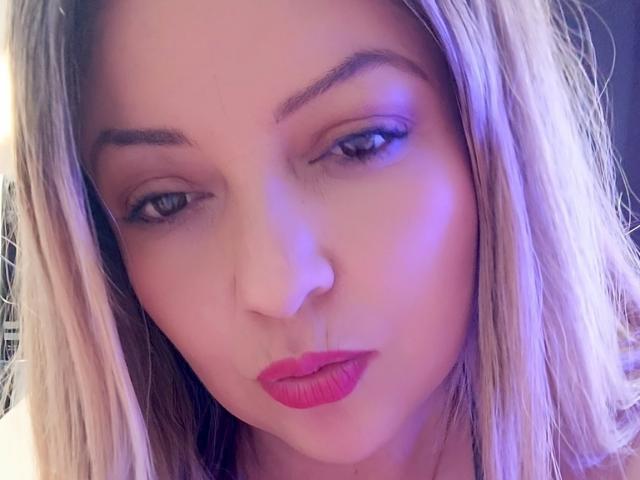 NatalyHami's face with blonde hair, pink lipstick, and blue eyeliner, looking down with a slight pout, in a soft purple light. Her tits are not visible.