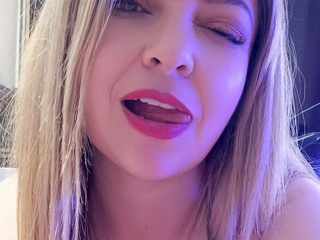 NatalyHami with blonde hair, winking, tongue out, red lipstick, and a slight smile, under purple lighting, showing a playful expression.
