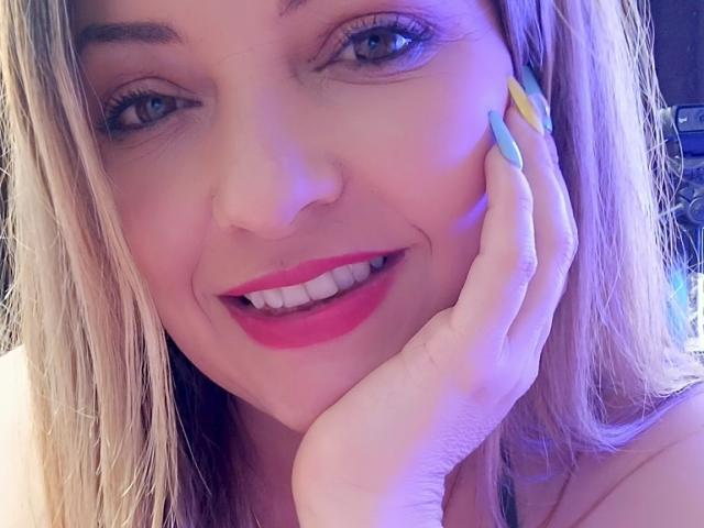 NatalyHami with blonde hair, blue eyes, and pink lipstick, smiling with her hand on her cheek, showing long, multicolored nails, and her white teeth. Her pink lips are slightly parted.