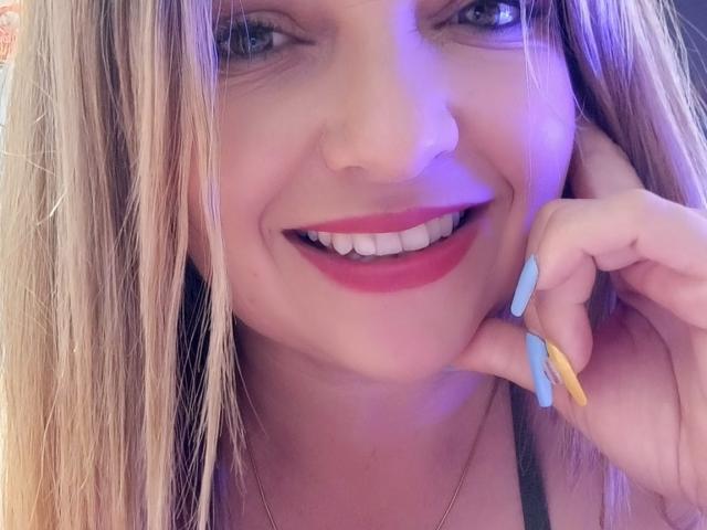 NatalyHami has blonde hair, blue eyes, pink lipstick, white teeth, and blue-yellow nails, smiling with her finger on her cheek. She's wearing a black top and a silver necklace.