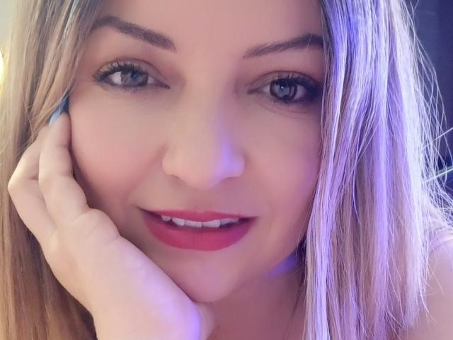 Il viso di NatalyHami, con i capelli biondi, gli occhi azzurri e il rossetto rosso, sorride leggermente, toccandosi la guancia con la mano, appare carina e sensuale. Il suo seno non è visibile..