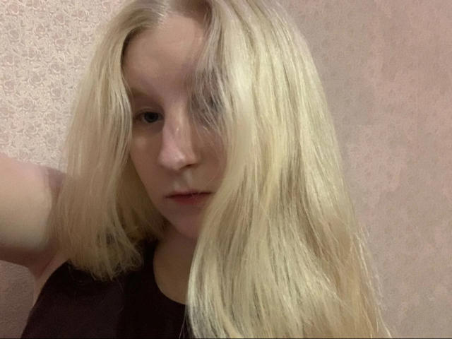 Blonde HyacinthBellin with long, straight hair covering one eye, wearing a dark top, has a neutral expression, pale skin, and a slightly parted mouth.
