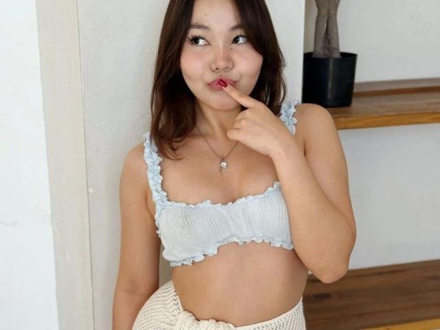 Sakura, con un reggiseno a fascia leggermente trasparente color azzurro, con un dito in bocca, un sorriso carino, un piccolo piercing al naso, capelli castani, pelle bianca, gonna lavorata a maglia, mensola di legno, pianta in vaso sullo sfondo..