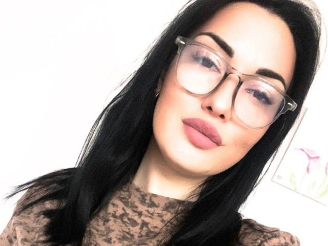 Xioni, con cabello negro, gafas y lápiz labial rosa, vestida con una blusa de encaje marrón, mirando ligeramente hacia un lado con una expresión neutra. Los senos no son visibles..