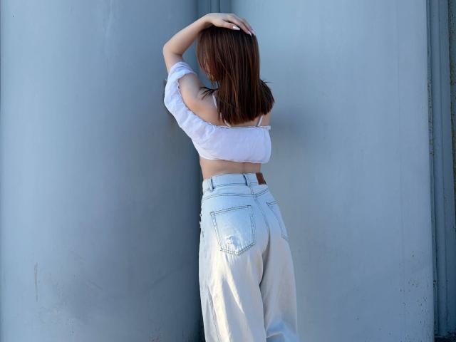 Donnetta Vannuland, avec ses cheveux bruns, son haut blanc à épaules dénudées et son jean blanc taille haute, les bras au-dessus de la tête, montrant ses fesses et un aperçu de ses seins contre un mur gris..