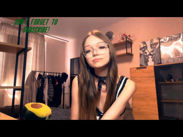 MeiWasinger with long brown hair, glasses, and a nose ring, wearing a black and white top, showing her tits, in a bedroom with clothes and furniture.