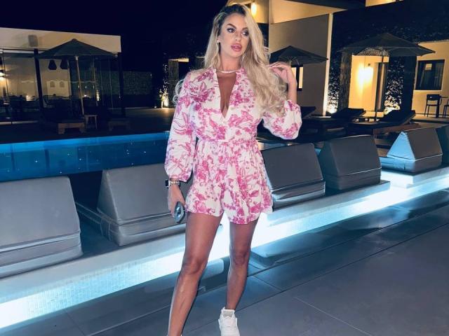 PearlPlenty with long blonde hair, pink floral dress, and white sneakers, standing by a blue-lit pool, holding a phone, looking at the camera with a confident smile.