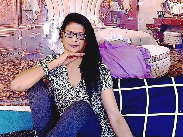 IndianAllure69 met lang zwart haar, een bril en een top met luipaardprint, zittend op een blauwe stoel, hand op kin, kijkend naar de camera, draagt donkerblauwe leggings, een paars kussen..