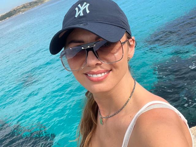ClarissaRose with tan skin, wearing a black NY cap, large sunglasses, white tank top, necklace, and smiling, has brown ponytail, blue ocean in background, looking sexy.