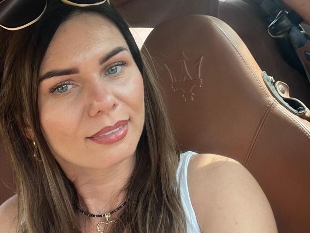 ClarissaRose ha gli occhi azzurri, i capelli castani, e sta tirando fuori la lingua seduta in un'auto con sedili in pelle marrone. Indossa una maglietta bianca e una collana nera..