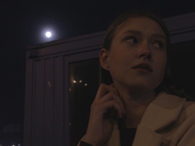 MaryGruen with a serious look, holding a phone to her ear, wearing a white coat, moon in dark night sky, right side of her face illuminated, looking up.