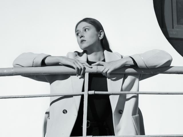 MaryaGruen with hands on railing, wearing a light coat, looking up, black top, medium tits, straight brown hair, white background, black and white photo.