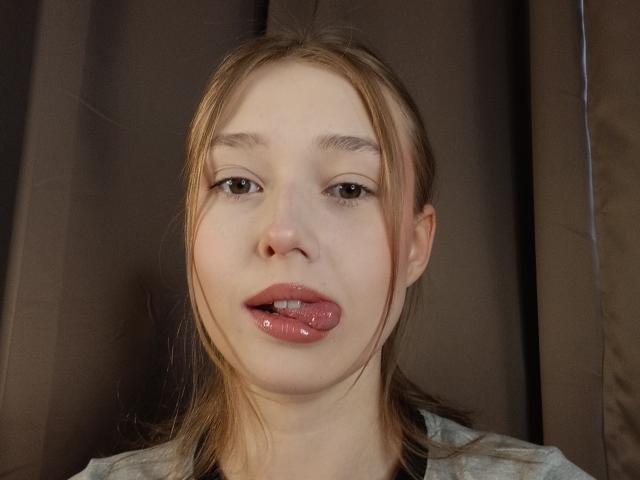 MaryaGruen with light brown hair, fair skin, and pink glossy lips, tongue slightly out, looking directly at the camera, brown curtain background.