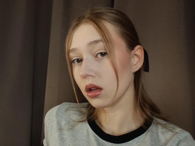 MaryaGruen has pale skin, brown eyes, and pouty pink lips with slight gloss. She's wearing a gray shirt with black trim, brown hair in a simple side-swept style.