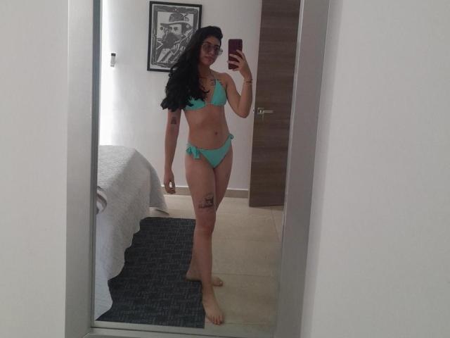 Spiegelfoto van MiluSky met lang zwart haar, die een turquoise bikini draagt, waarbij haar borsten en billen zichtbaar zijn, met een tatoeage op haar rechter dij, en die een rode telefoon vasthoudt..