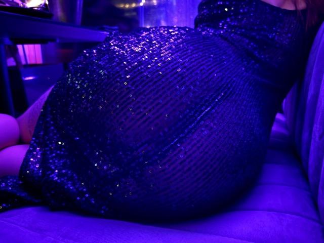 El trasero de MiraGi aparece prominentemente en una toma, vistiendo un vestido brillante, con lentejuelas, de color púrpura oscuro, y sentada en un sofá iluminado con luz púrpura. Se puede apreciar la textura del vestido..