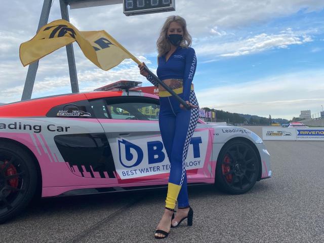 Atomblondy-hot stands in front of a racing car, holding a yellow flag, wearing a blue and yellow checkered jumpsuit, black heels, and a mask, with tattoos visible.