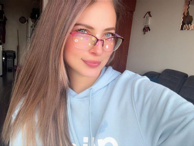 AmbeerWood, com cabelos castanhos longos, olhos azuis e óculos florais rosa, vestindo um moletom azul claro, sorrindo levemente. Seus pequenos seios estão parcialmente visíveis. Luz natural suave..