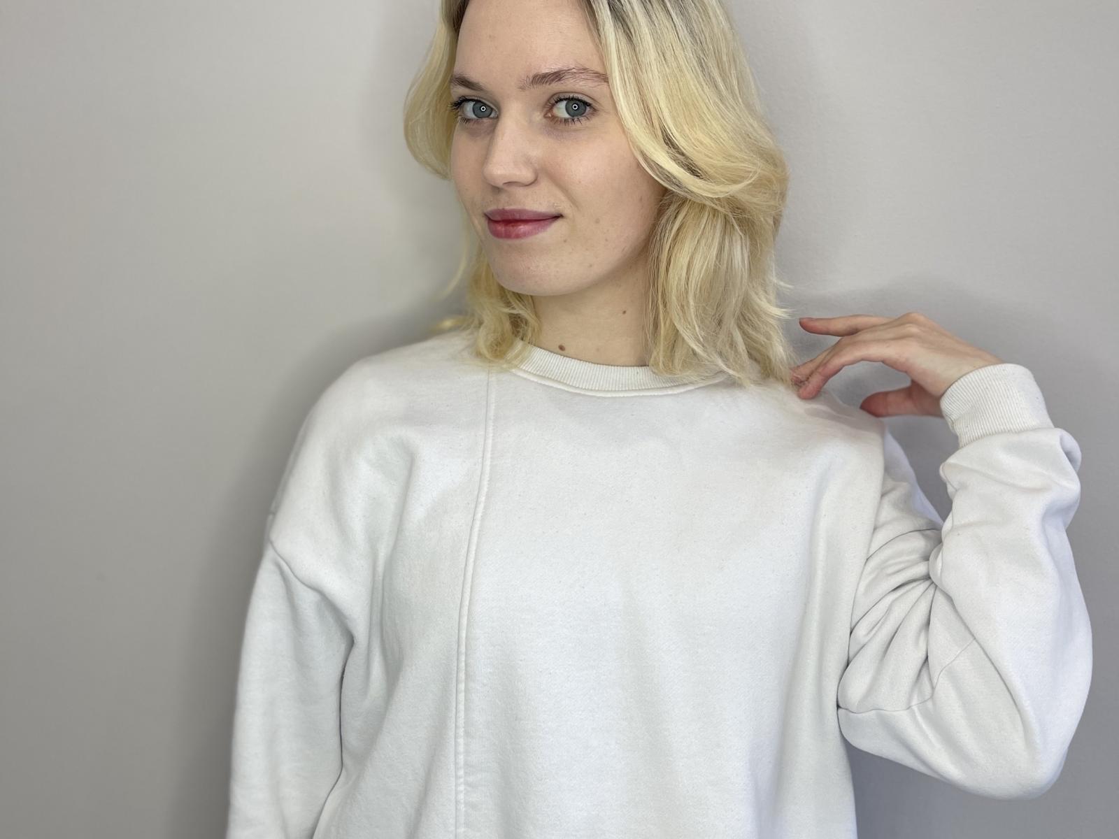 SheraSwayzer with blonde hair, wearing a white sweatshirt, hand near head, smiling slightly, red lipstick, blue eyes, soft light, casual, relaxed, upper body, white wall background.