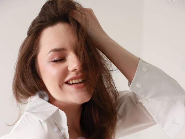 Dreia with brown hair, white shirt, eyes closed, smiling, hand in hair, showing her teeth, soft light, slightly messy hair, relaxed, casual, attractive, cute.