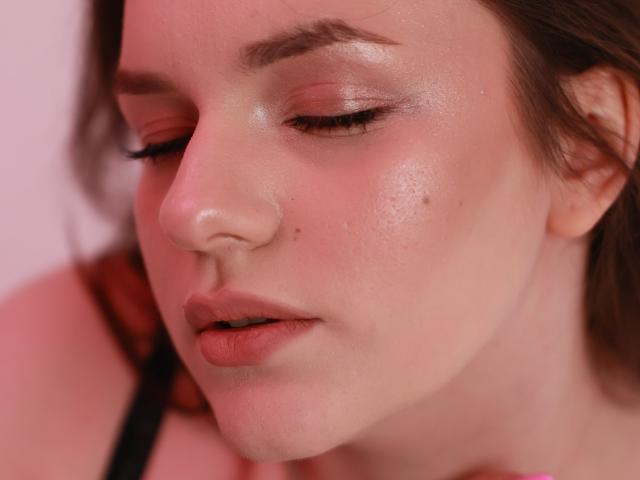 Le visage de Dreia, avec sa peau rosée, ses yeux fermés et son maquillage discret, présente une petite tache de beauté sur sa joue gauche. Ses lèvres sont légèrement entrouvertes, et elle porte un bandeau noir..